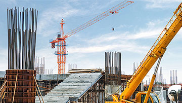 Construction site with steel columns, crane, and yellow excavator under blue sky.
