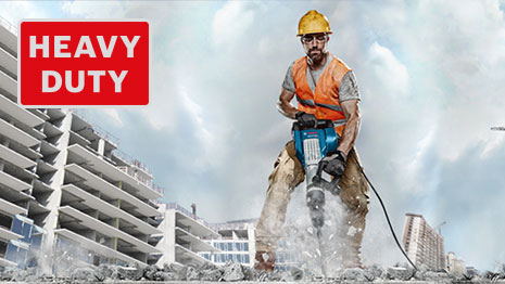 A worker with a jackhammer in a construction area, surrounded by buildings.