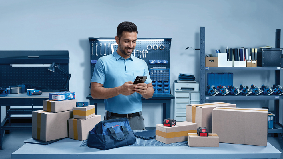 A man in a blue polo shirt stands in a workshop, looking at his smartphone. Around him are boxes and tools.