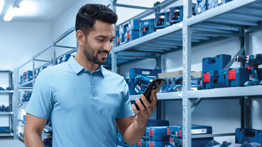 A man in a light blue polo shirt stands in a room full of Bosch Professional toolboxes and looks at his smartphone.
