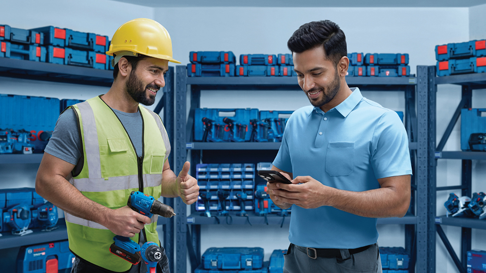 A smiling worker in a yellow helmet and green vest gives a thumbs up while talking to a man in a blue polo shirt, surrounded by blue toolboxes.
