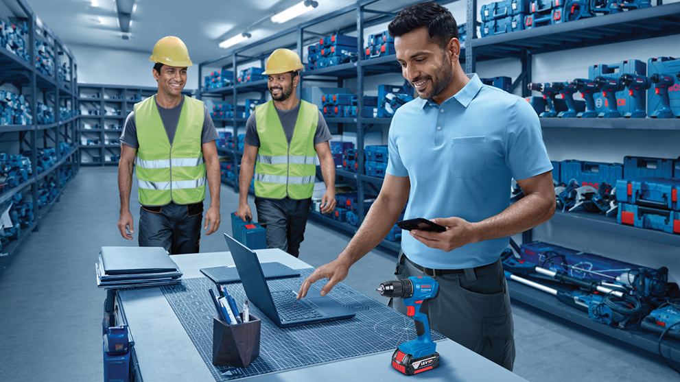 A man in a blue polo shirt stands at a table with a laptop and a Bosch Professional cordless screwdriver. In the background, two men wearing yellow helmets and safety vests are in a well-organized tool storage.