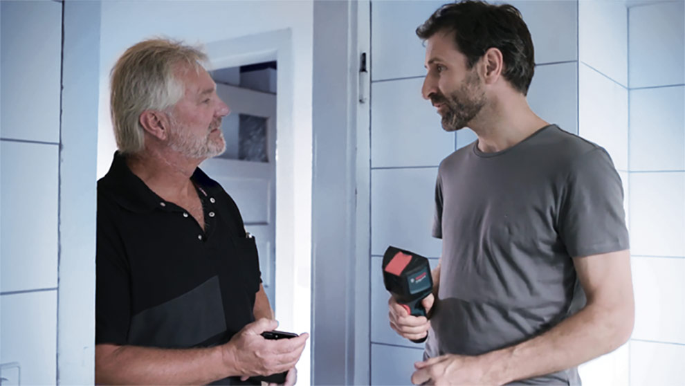 Two men stand in a bright room, one holding a Bosch tool.
