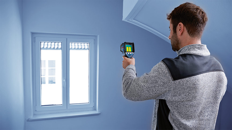 A man with an infrared thermometer stands in front of a window.